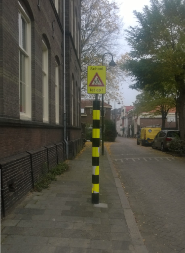 Foto met daarop een hoge regenboogpaal met zwarte en gele strepen en daar bovenop een waarschuwingsbord met de tekst: school, let op!