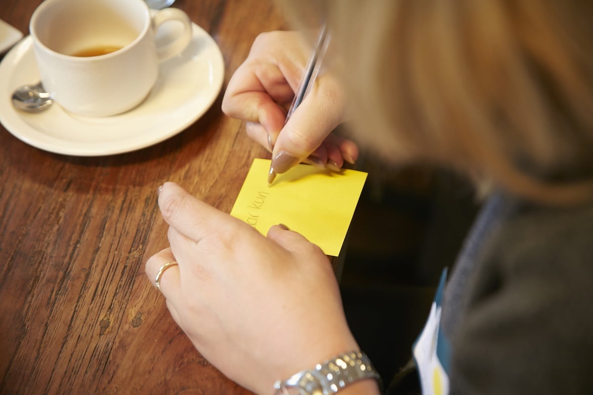 Een vrouw schrijft op een geel notitieblok, met daarnaast een lege kop koffie op de tafel.