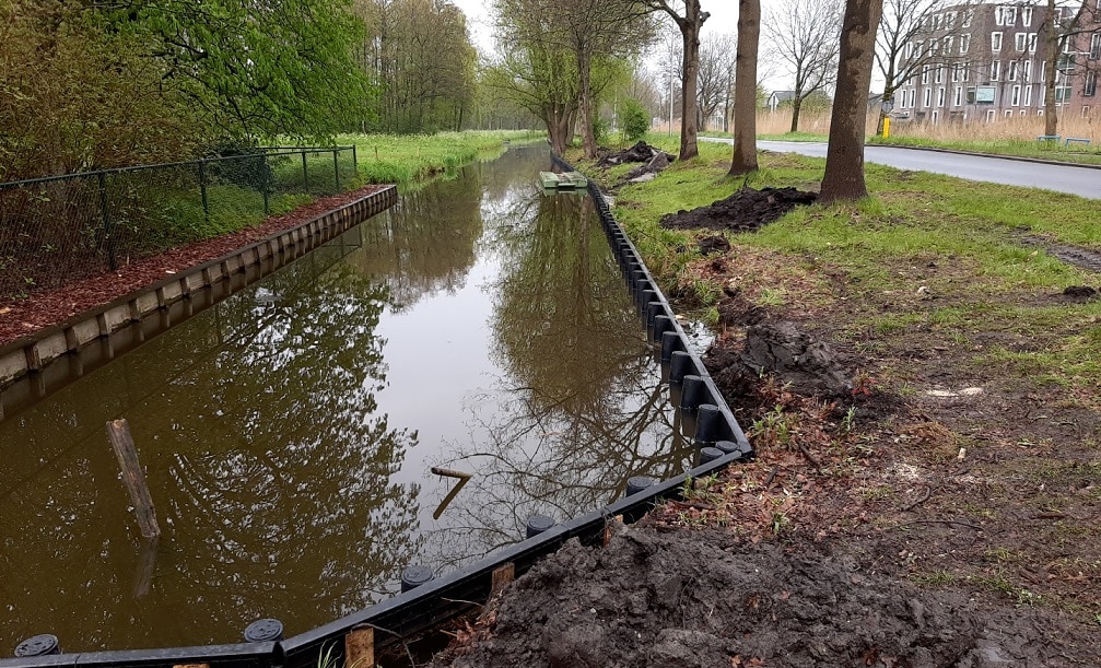 Een voorbeeld van beschoeiing. Een sloot langs de weg, met houten paaltjes en planken om de oever te beschermen. Bomen staan aan weerszijden van de sloot, in de verte een woonwijk.