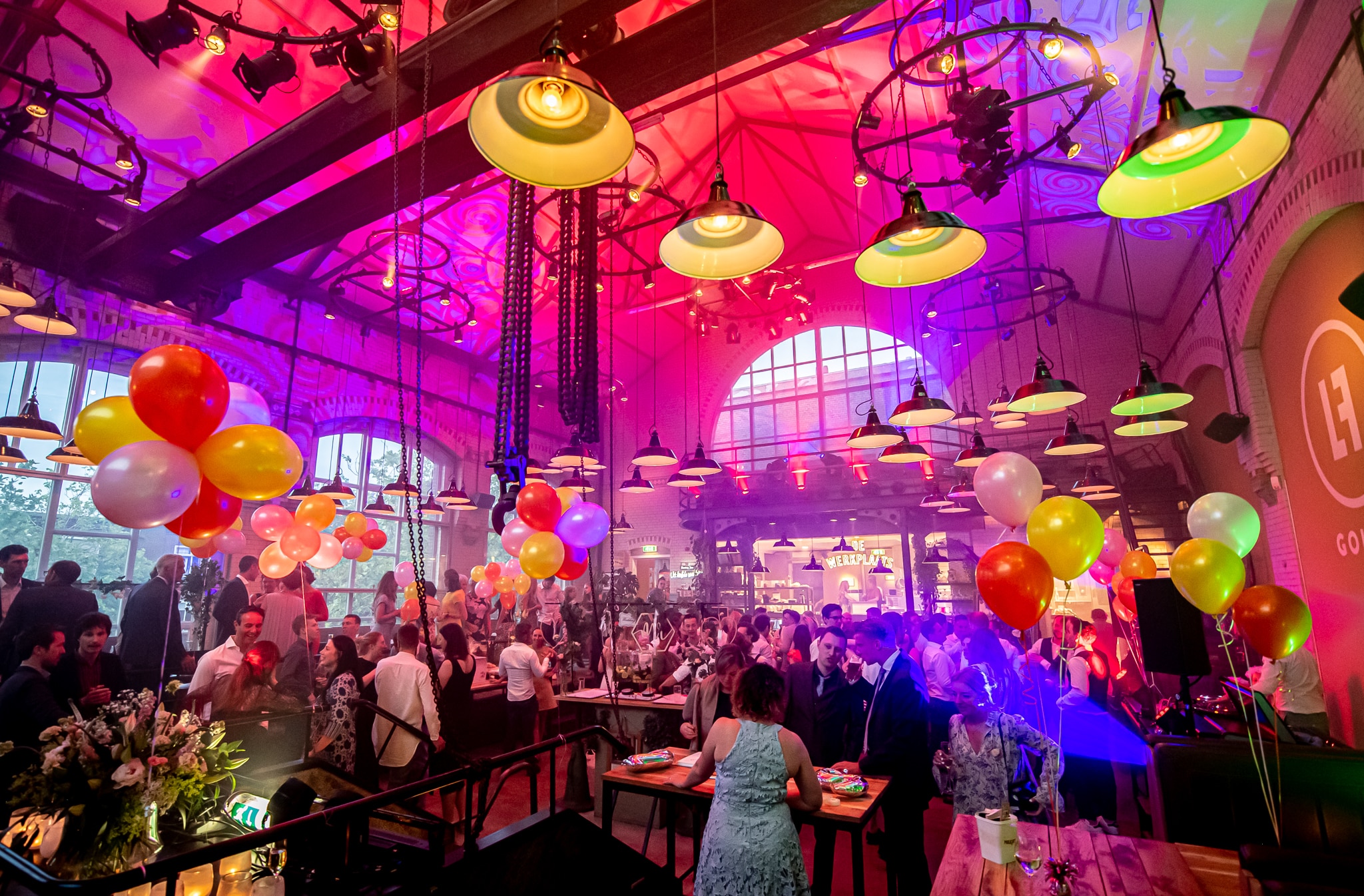 Op de foto is de binnenkant van de Lichtfabriek in Gouda te zien. De zaal is gevuld met mensen en er hangen veel ballonnen.