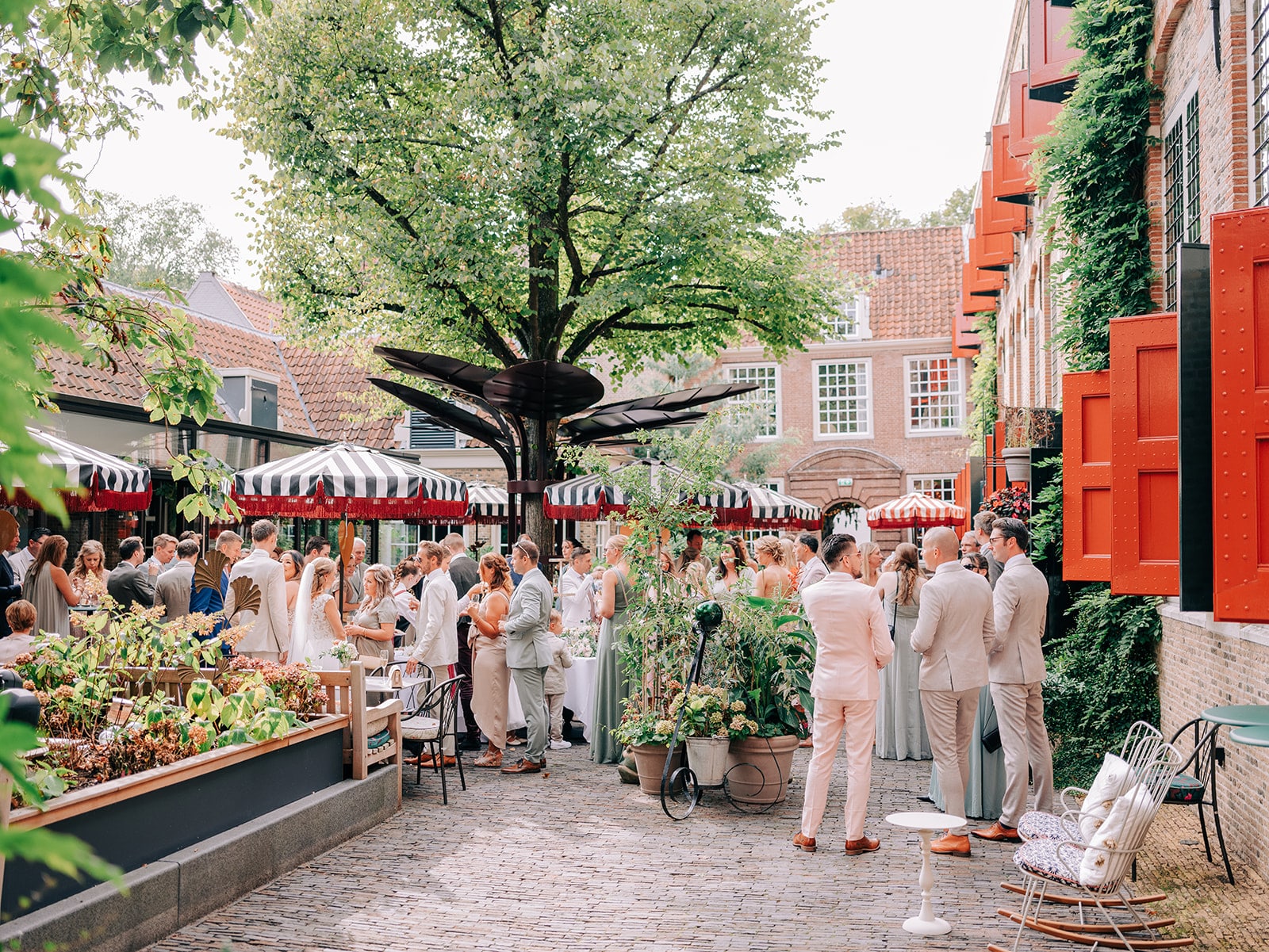 Op de foto is de buitenplaats van het Weeshuis Gouda te zien, gevuld met bruiloftsgasten. In het midden staat een grote boom en er staat een aantal parasols.
