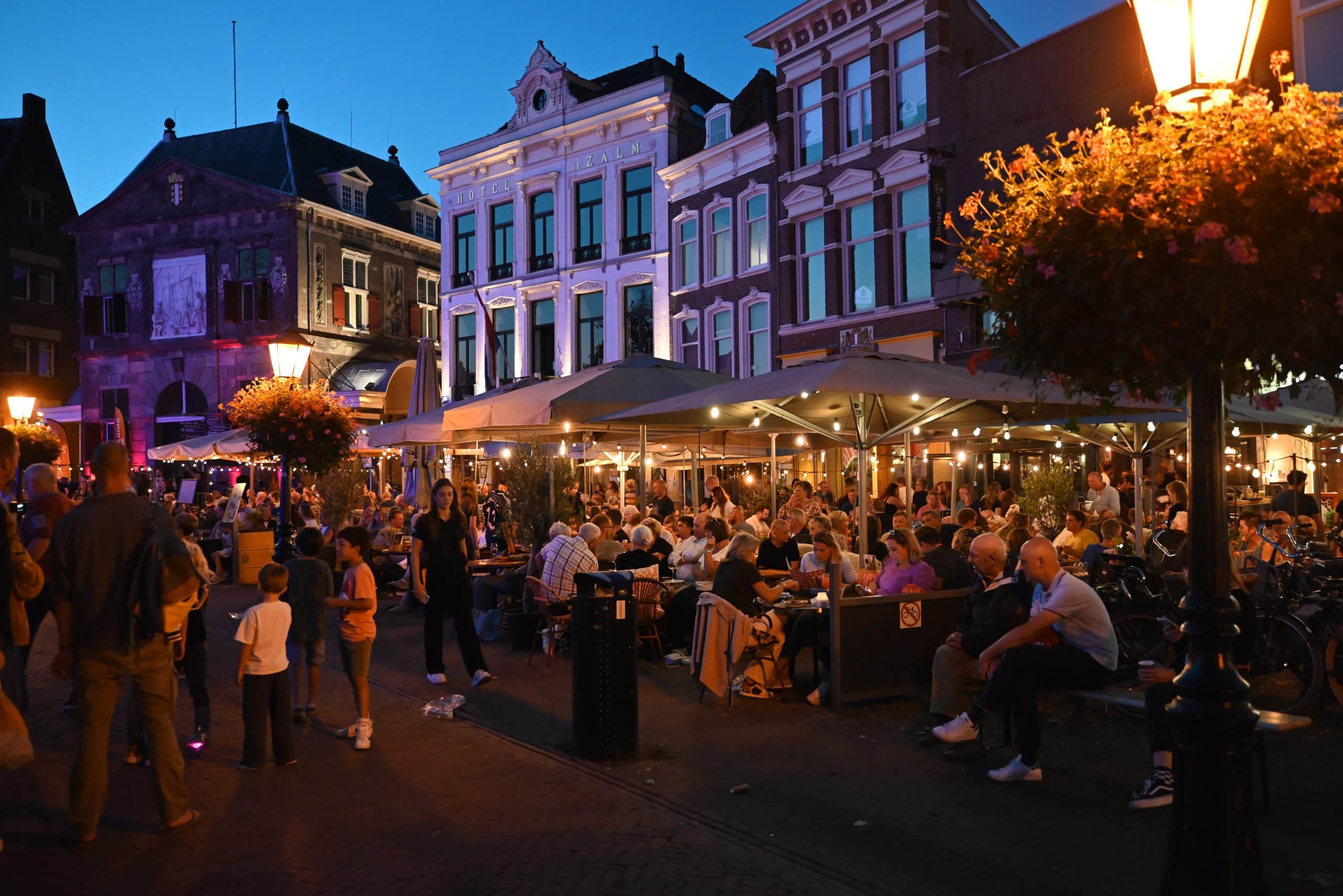 Op de foto staan terrassen op de Markt. Het is avond en de terrassen zitten vol met mensen.