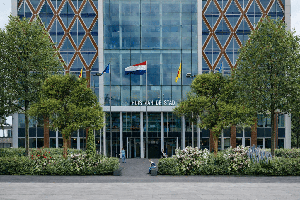 Op de afbeelding is het vooraanzicht van het Huis van de Stad te zien. Er zijn verschillende bomen en beplanting te zien, die in de aankomende periode worden aangeplant. De beplanting is voornamelijk groen met een aantal kleuraccenten. Er is ook een aantal zitranden ingetekend. In de bestaande situatie is het plein versteend. De gemeente vergroent het plein