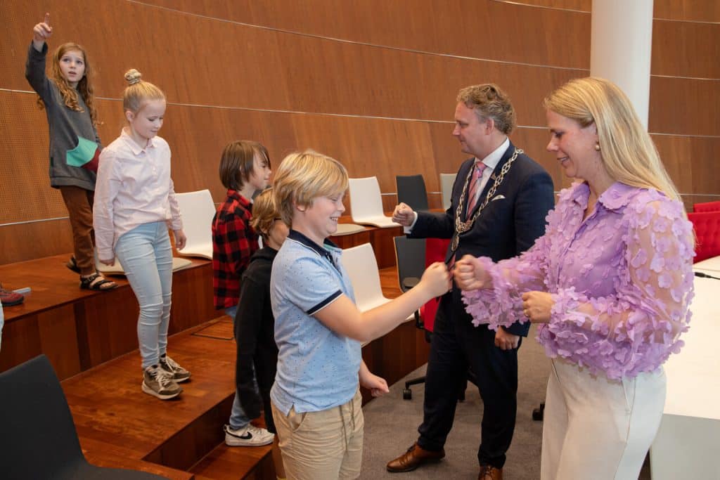 Burgemeester Pieter Verhoeve en wethouder Anna van Popering-Kalkman in de raadszaal met een aantal kinderen. De kinderen geven de burgemeester en de wethouder een boks.